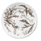 A round ceramic plate with a brown seaweed pattern on an off-white background, displayed on a rustic dark wood plank surface.