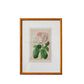 A framed botanical art print of a pink rose with green leaves, set in a gold frame and leaning against a textured, distressed white plaster wall.