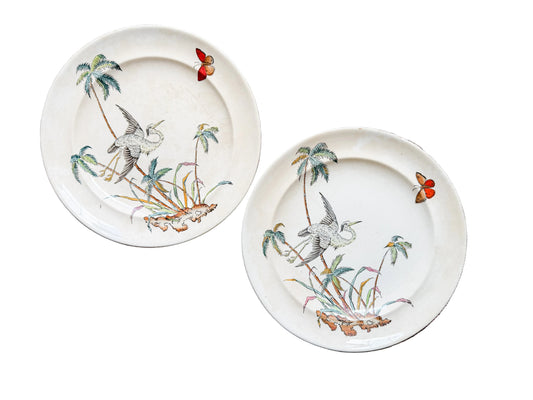 Two round ceramic plates with a vintage hand-painted design of a bird, palm trees, and butterflies, displayed on a rustic wooden background.