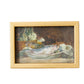 A framed still life oil painting depicting a fish on a plate, housed in a light natural wood frame and leaning against a textured off-white plaster wall.