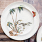 A white ceramic plate with a vintage-style tropical and bird illustration, featuring palm trees, a bird in flight, a butterfly, and a leopard, displayed on a rustic wooden surface.