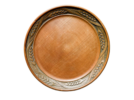 A round wooden serving platter with a smooth, polished brown center and a carved decorative border featuring wheat stalks, shown from a top-down view on a light gray textured background.