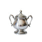 A polished silver lidded sugar bowl with ornate handles and a decorative pedestal base, photographed on a white marble surface against a matching marble background.