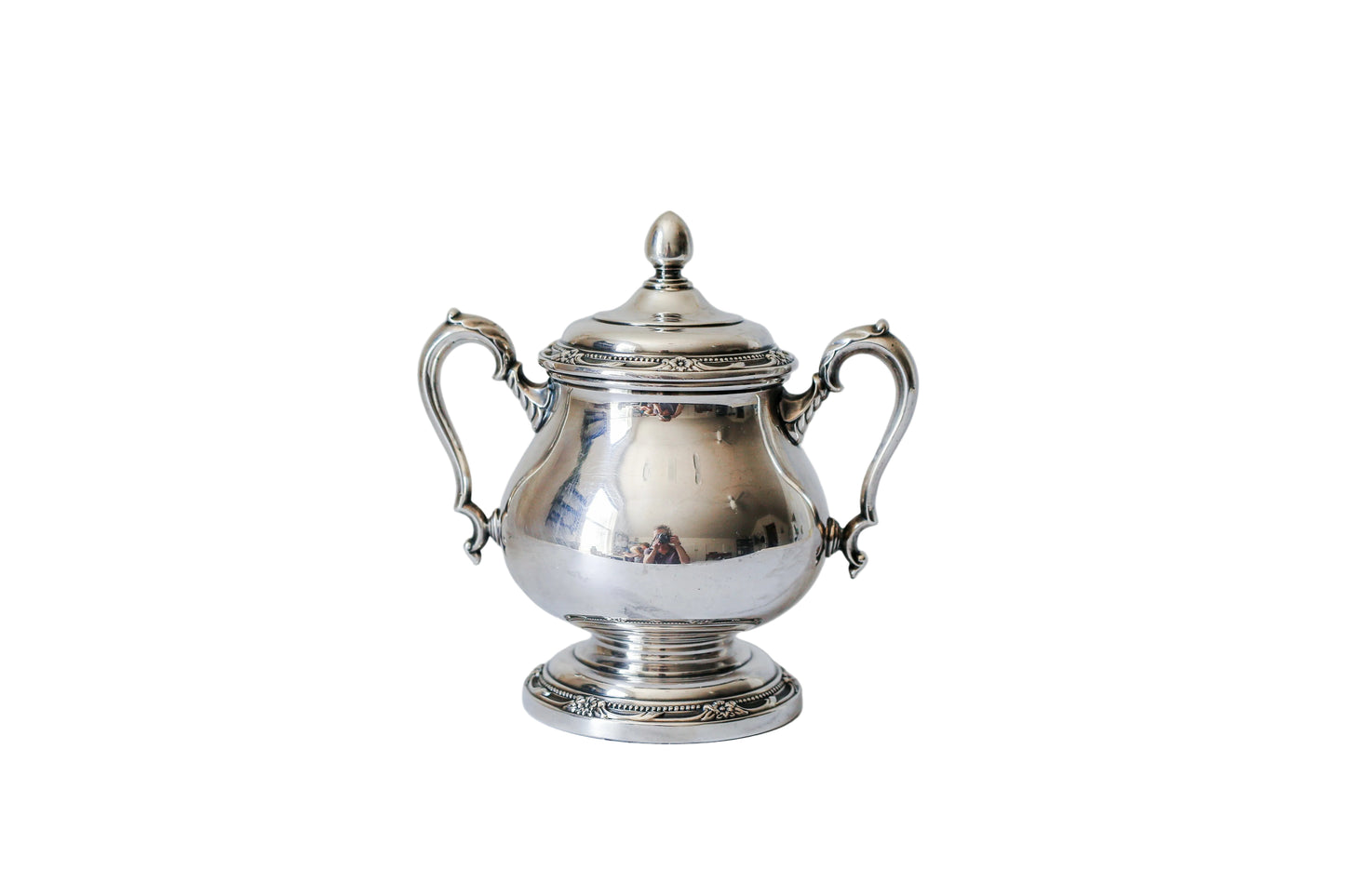 A polished silver lidded sugar bowl with ornate handles and a decorative pedestal base, photographed on a white marble surface against a matching marble background.