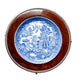 A round decorative wall plate featuring a blue and white illustrated porcelain design in the center, surrounded by a polished reddish-brown wooden ring and a silver-toned metal rim, displayed on a light textured fabric background.