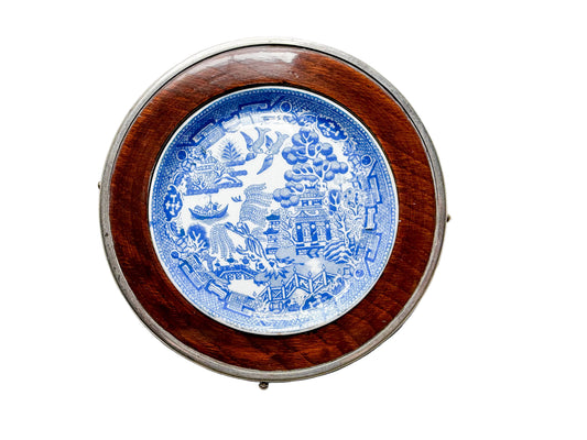 A round decorative wall plate featuring a blue and white illustrated porcelain design in the center, surrounded by a polished reddish-brown wooden ring and a silver-toned metal rim, displayed on a light textured fabric background.