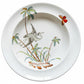 A white ceramic plate featuring a hand-painted illustration of a heron, palm trees, and a butterfly, displayed on a rustic wooden background.