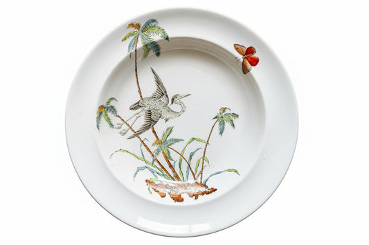 A white ceramic plate featuring a hand-painted illustration of a heron, palm trees, and a butterfly, displayed on a rustic wooden background.