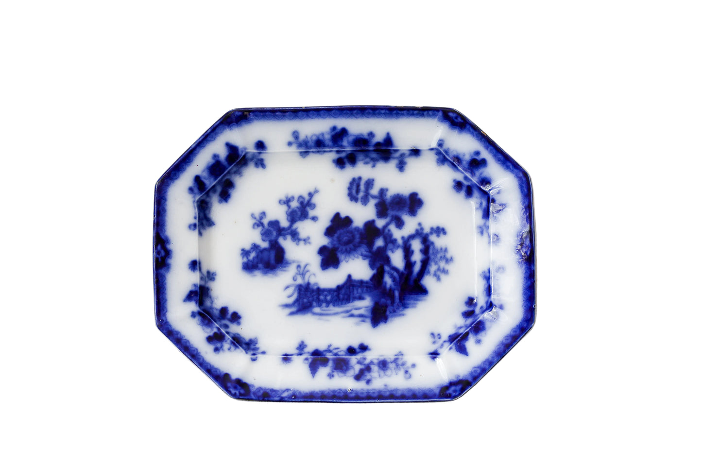 An octagonal white porcelain serving platter with blue hand-painted floral and landscape motifs, set against a textured, distressed off-white wall.