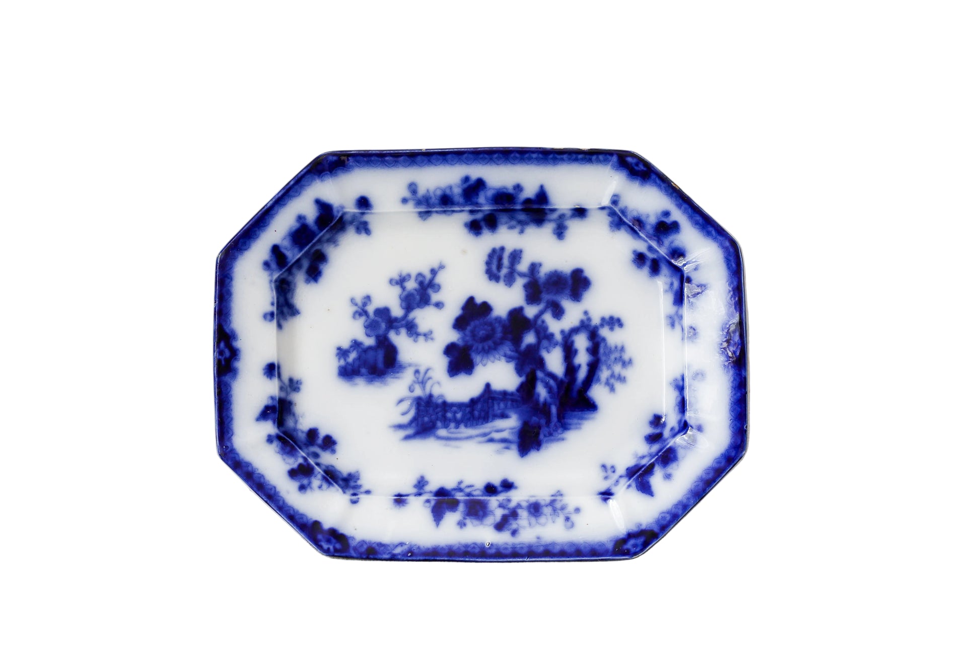 An octagonal white porcelain serving platter with blue hand-painted floral and landscape motifs, set against a textured, distressed off-white wall.