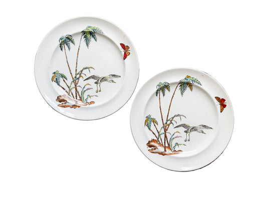 Two white ceramic plates with hand-painted tropical designs, including palm trees, birds, and butterflies, displayed on a rustic wooden plank background.
