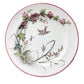 A round white ceramic plate with a red rim and a botanical illustration featuring pink flowers, green leaves, and a small bird, displayed on a rustic wooden plank surface.