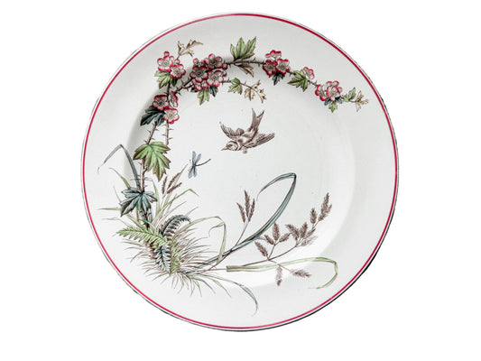 A round white ceramic plate with a red rim and a botanical illustration featuring pink flowers, green leaves, and a small bird, displayed on a rustic wooden plank surface.