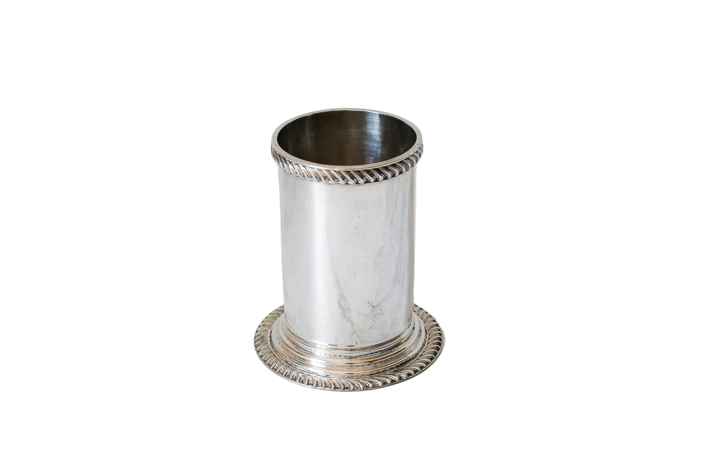 A small silver cylindrical holder or cup with decorative twisted rope-style trim at the rim and base, standing on a white marble surface against a white marble background.