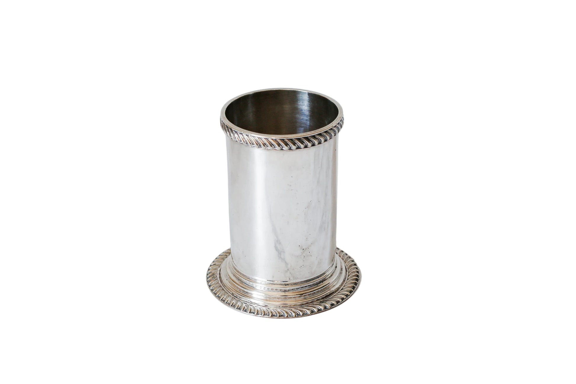 A small silver cylindrical holder or cup with decorative twisted rope-style trim at the rim and base, standing on a white marble surface against a white marble background.