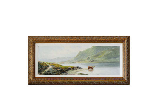 A framed landscape painting of a coastal scene with green hills, water, and a cow on the beach, set in an ornate gold frame against a textured white wall.